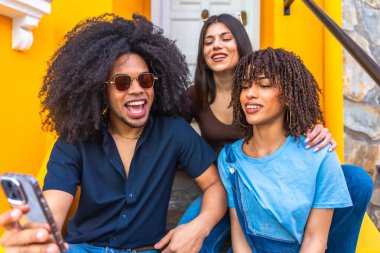 Üç mutlu genç arkadaş renkli bir binanın merdivenlerinde otururken akıllı telefonlarıyla selfie çekiyorlar.