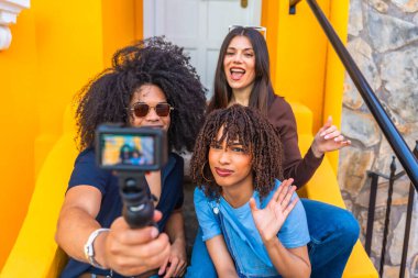 Canlı bir video blogunu renkli arka planlara sahip bir şehir ortamında çekerken kameraya el sallayan ve gülümseyen üç etkili kişi