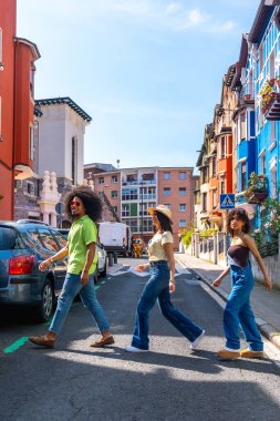 Üç arkadaş neşeli bir mahallede yürüyüş yapıyorlar, güneşli bir günde karşıdan karşıya geçiyorlar.