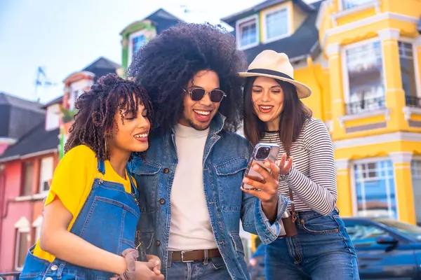 Üç neşeli genç turist gülümseyip akıllı bir telefon kullanırken renkli ve hayat dolu canlı bir şehir sokağının keyfini çıkarıyorlar.