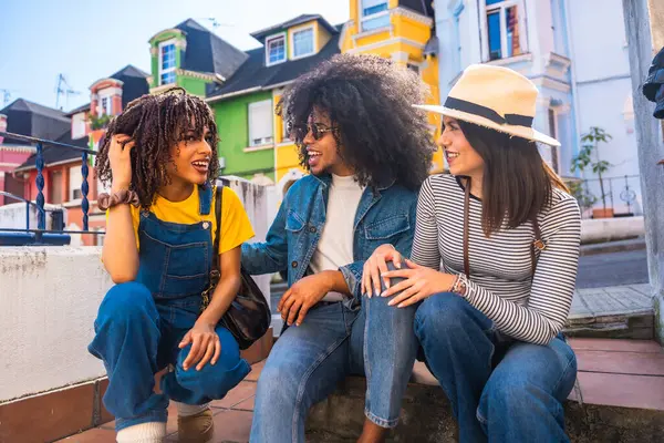 Üç genç turist renkli bir caddenin merdivenlerinde oturmuş sohbet ediyor ve yaz tatillerinin tadını çıkarıyorlar.