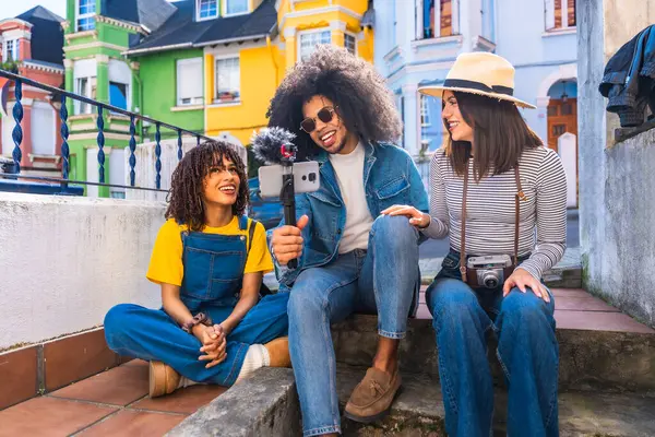 Gülümseyen arkadaşlar renkli bir mahallede mikrofonlu akıllı telefon kullanarak video bloğu oluşturuyorlar.