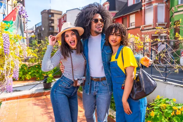 Üç genç turist ellerinde kamerayla sarılıp gülümsüyor, renkli bir şehir ortamında tatilin tadını çıkarıyorlar.