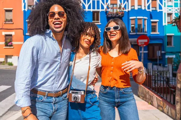 Güneşli bir yaz tatili boyunca canlı, renkli bir sokakta birlikte yürüyen ve gülümseyen üç neşeli genç turist.