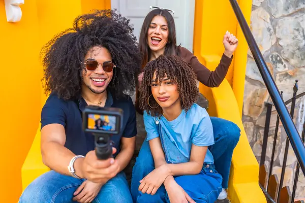 Üç mutlu çoklu etnik video kaydedicisi açık havada renkli sarı merdivenlerde oturarak bir video blogu kaydediyor.