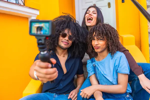 Üç Latin arkadaş bir video bloğu çekiyorlar. Titreşimli sarı basamaklarda otururken gülüp eğleniyorlar.