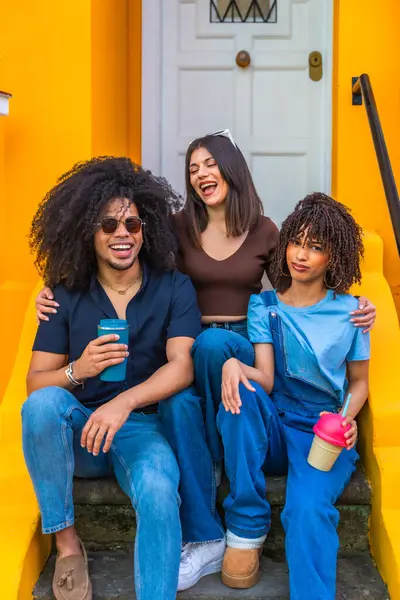 Üç genç arkadaş birlikte rahat vakit geçiriyorlar, merdivenlerde oturup gülüyorlar ve ellerinde yeniden kullanılabilir bardaklar tutuyorlar.