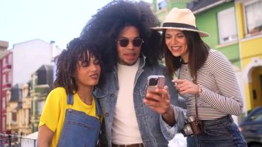 Üç neşeli arkadaş, iki kadın ve bir adam, akıllı bir telefona bakıyorlar ve bir şehir gezisi sırasında birlikte gülüyorlar.