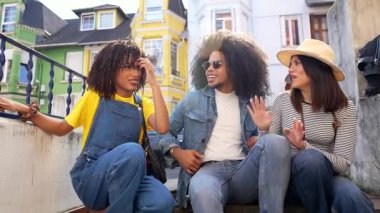 Üç arkadaş, günlük yaz kıyafetleri içinde, canlı bir sohbete dalarken canlı ve renkli bir şehirde oturuyorlar.
