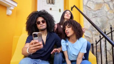 Genç arkadaşlar parlak sarı şehir basamaklarında otururken selfie çekiyorlar, neşeli bir arkadaşlık anı yakalıyorlar ve akıllı telefonlarla bağlantı kuruyorlar.