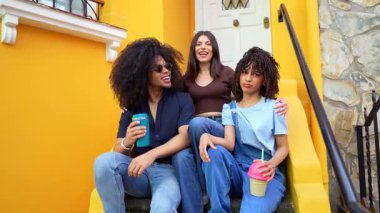 Üç arkadaş, canlı sarı bir binanın merdivenlerinde otururken gülüp sohbet ediyorlar. İçeceklerin tadını çıkarıyorlar.