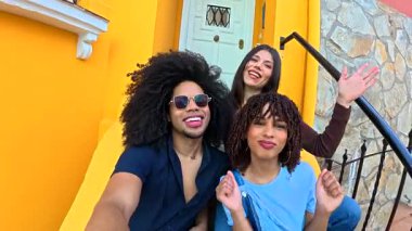 Canlı sarı bir binanın merdivenlerinde fotoğraf çekerken üç neşeli arkadaş poz veriyor ve komik surat ifadeleri takınıyor.