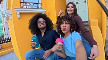 Üç arkadaş, iki kadın ve bir erkek, canlı sarı bir duvarın önünde içki içerken selfie çekiyorlar.