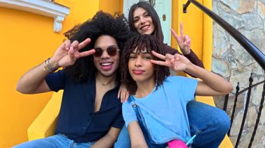 Neşeli çoklu etnik arkadaşlar içeceklerle kaynaşıyor, canlı sarı basamaklarda oturuyor, pozitif enerji yayıyor ve yaz havası yayıyor.