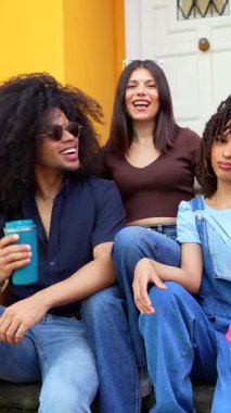 Üç neşeli arkadaş, iki kadın ve bir erkek merdivenlerde oturup gülüyor, içiyor ve birbirlerinin arkadaşlığından keyif alıyorlar.