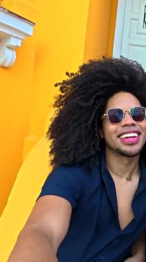 Üç neşeli genç arkadaş, iki kadın ve bir adam, parlak sarı bir duvarın önünde poz veriyorlar ve komik suratlar yapıyorlar.