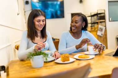 Kahve molasından zevk alan iki genç kadın, doğal ışıkla dolu canlı, modern bir kafede sohbet edip gülüyorlar.