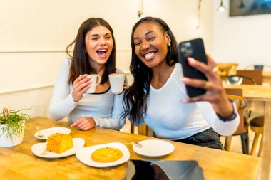 İki genç kadın bir kafede kahve içip hamur işi yerken akıllı telefonlarıyla selfie çekiyorlar ve birlikte vakit geçiriyorlar.
