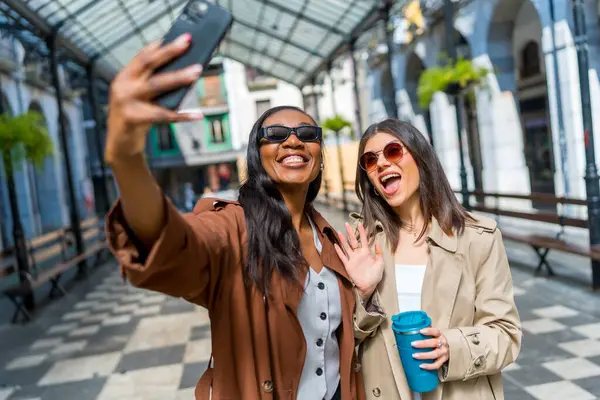 İki neşeli iş kadını akıllı bir telefonla selfie çekiyor ve şehirde yürürken kameraya el sallıyorlar.