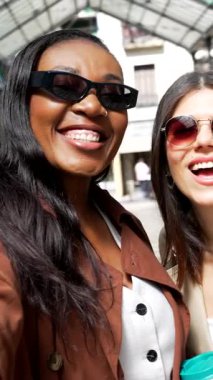 Kadın arkadaşlar birlikte poz veriyor, güneş gözlüğü takarken selfie çekiyor güneşli bir günde kentsel mimari arka planla kaplı bir yürüyüş yolunun altında dikiliyorlar.