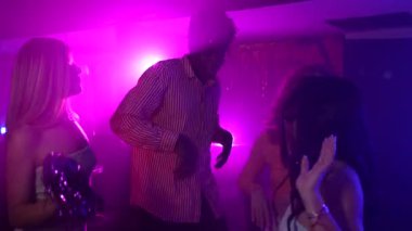 Canlı bir gece kulübü sahnesi. Dans eden, müzik paylaşan, renkli ışıklar ve canlı bir atmosfer arasında enerjik bir coşkuyla kutlama yapan genç arkadaşlar.