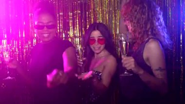 Güneş gözlüğü takan ve şampanya bardakları tutan genç kadınlar altın renkli perdeli bir fotoğraf kabininde eğleniyorlar.