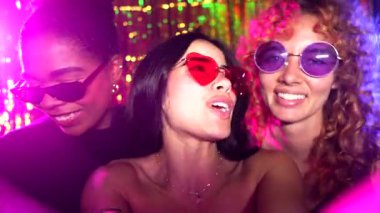 Renkli güneş gözlüklü genç kadınlar enerjik disko partisinde unutulmaz anlar yakalıyorlar, canlı bir gece hayatı kutlaması yaşarken arkadaşlarıyla neşeli selfie çekiyorlar.