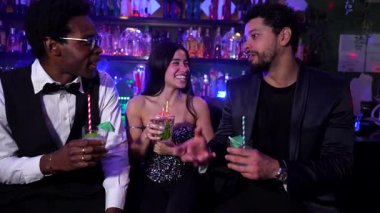 Üç arkadaş, iki erkek ve bir kadın, bir gece kulübünde kokteyl içerken gülüp sohbet ediyorlar.