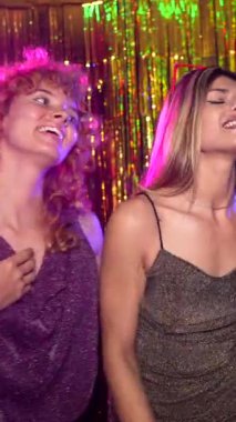 Kadın arkadaşlar birlikte şarkı söyleyip dans ediyorlar. Karaoke parti atmosferinde canlı bir enerji yaratıyorlar. Altın arka plan ve renkli ışıklar.