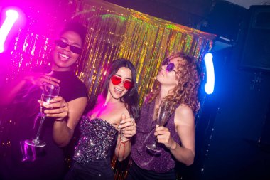 Üç kadın ellerinde şampanya bardakları ve renkli ışıkları ve altın bir perdesi olan bir gece kulübünde eğleniyorlar.