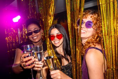 Üç çok ırklı bayan arkadaş bir gece kulübünde şampanya bardaklarıyla kadeh kaldırıyor, gece hayatının tadını çıkarıyor ve renkli güneş gözlüğü takıyorlar.