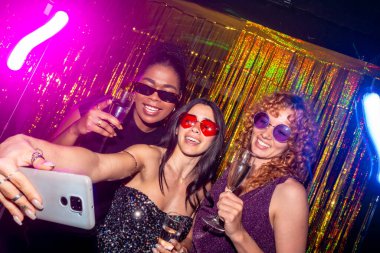 Üç çok ırklı bayan arkadaş bir gece kulübünde köpüklü şarap içerken selfie çekiyorlar. Neon ışıkları ve altın folyo perdesiyle kutlama yapıyorlar.