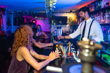 Modaya uygun bir gece kulübünde canlı kokteyller hazırlayan barmen, canlı müzik ve renkli ışıkların ortasında hareketli bar tezgahında genç kadınlara servis yapıyor.