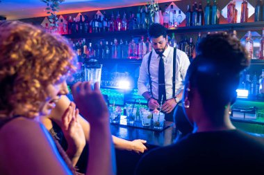 Profesyonel barmen gece kulübünde neşeli bayan müşterilere kokteyller hazırlıyor, içkileri karıştırıyor ve canlı bir ortam yaratıyor.