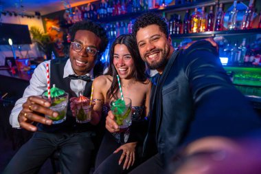 Üç arkadaş gülümseyip selfie çekerken canlı bir barda renkli kokteyllerin tadını çıkarıyorlar. Arkadaşlık ve eğlenceyi kutluyorlar.