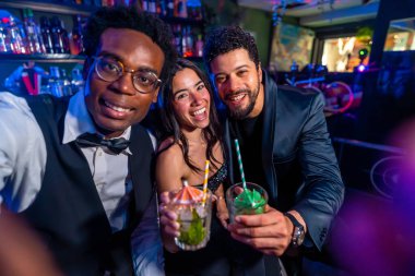 Neşeli çoklu etnik arkadaşlar selfie çekiyorlar ve gece kulübünde eğlenirken kokteyl gösteriyorlar.