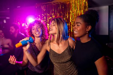 Üç genç kadın canlı bir karaoke gecesinin tadını çıkarıyorlar. Renkli ışıkları ve arka planda parıldayan süslemeleri olan bir mikrofona şarkı söylüyorlar.