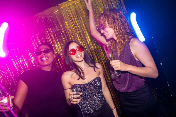 Şampanyalı, dans eden ve neon ışıkları ve pırıl pırıl altın arkaplanı olan şık bir gece kulübünde eğlenen zarif genç kadınlar.