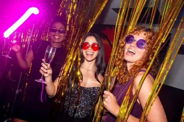 Renkli güneş gözlüklü üç neşeli genç kadın ellerinde köpüklü şarap bardakları, altın süslemeleri ve neon ışıklarıyla canlı bir disko partisinin tadını çıkarıyorlar.