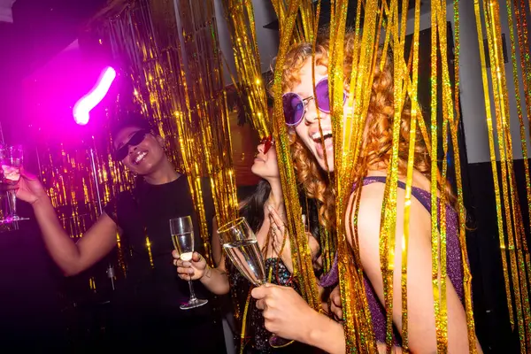 Genç kadınlar canlı bir disko partisinden zevk alıyorlar, ellerinde köpüklü şarap bardakları ve parıldayan altın perdelerin arkasında poz veriyorlar.
