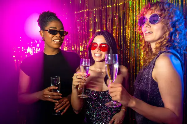 Üç çok ırklı bayan arkadaş canlı bir gece kulübünde renkli ışıklar ve parıltılı dekorasyonlar arasında içki içiyorlar.