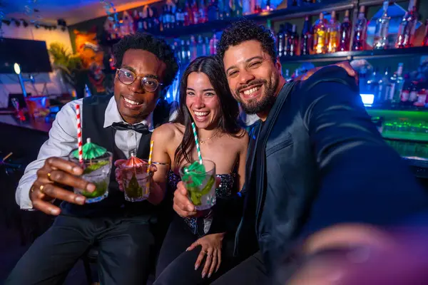 Üç arkadaş gülümseyip selfie çekerken canlı bir barda renkli kokteyllerin tadını çıkarıyorlar. Arkadaşlık ve eğlenceyi kutluyorlar.