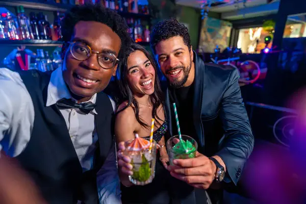 Neşeli çoklu etnik arkadaşlar selfie çekiyorlar ve gece kulübünde eğlenirken kokteyl gösteriyorlar.