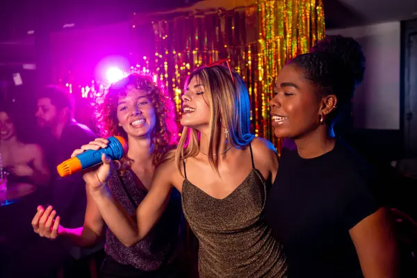 Üç genç kadın canlı bir karaoke gecesinin tadını çıkarıyorlar. Renkli ışıkları ve arka planda parıldayan süslemeleri olan bir mikrofona şarkı söylüyorlar.