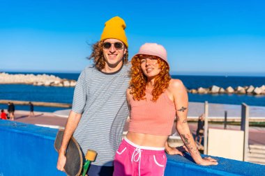 Deniz kenarında kaykaylarıyla poz veren iki genç kaykaycı sevgi ve kahkahalarla dolu güneşli bir yaz gününün tadını çıkarıyorlar.