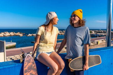 İki genç patenci deniz kenarındaki mavi bir duvarda oturmuş, uzun kaykaylarını tutuyor ve güneşli bir günün tadını çıkarıyorlar.