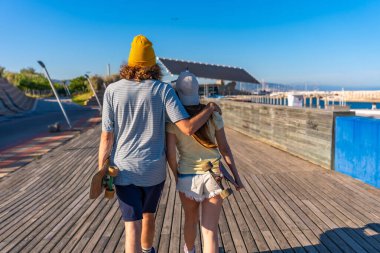 Genç çift güneşli bir tahta yolda el ele yürüyor, kaykay taşıyor ve okyanusun kenarında kaygısız bir günün tadını çıkarıyor.