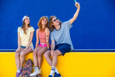 Üç neşeli arkadaş, mavi arka planı olan sarı bir duvarda otururken akıllı bir telefonla selfie çekiyorlar.