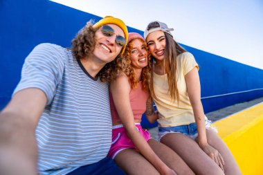 Üç genç arkadaş gülümsüyor ve canlı mavi ve sarı bir duvarın önünde selfie çekiyorlar.