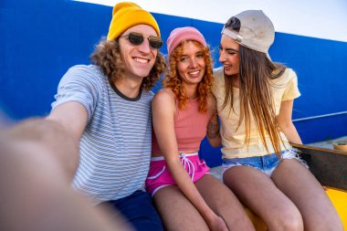 Üç genç arkadaş gülümsüyor ve dışarıda eğleniyorlar, gündelik tarzda kucaklaşıyorlar ve açık mavi gökyüzünün altında güneşli bir günün tadını çıkarıyorlar.